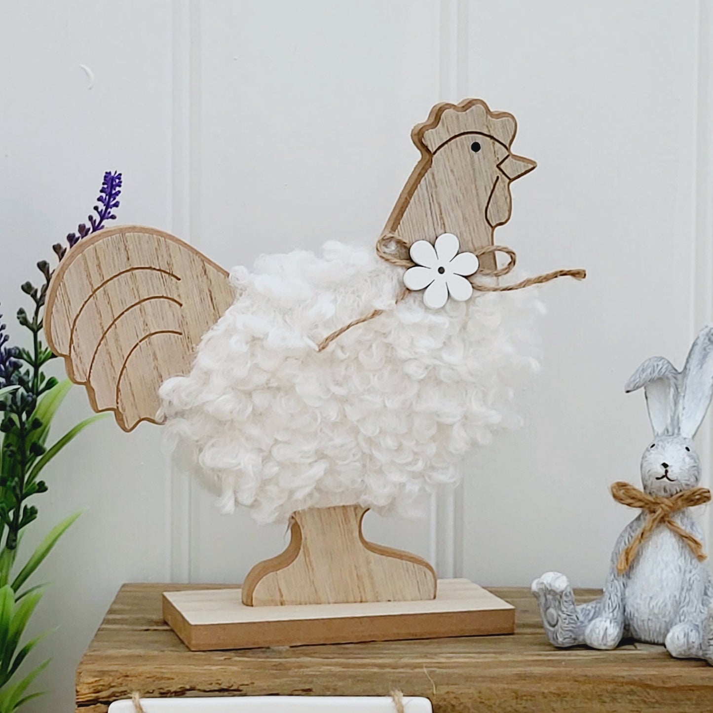 A freestanding wooden chicken ornament with a white fluffy body and a jute flower bow around neck, displayed on a wooden bench next to a grey rabbit ornament