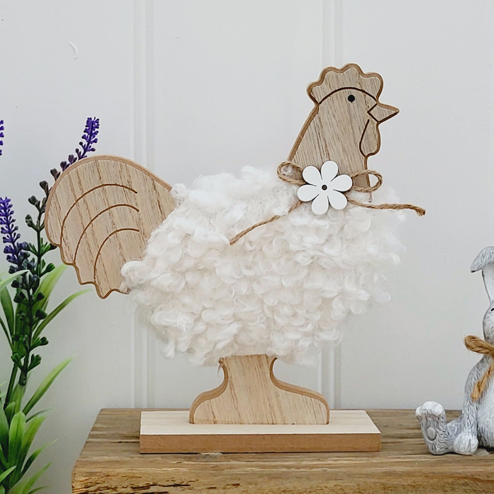 A freestanding wooden chicken ornament with a white fluffy body and a jute flower bow around neck, displayed on a wooden bench next to a grey rabbit ornament