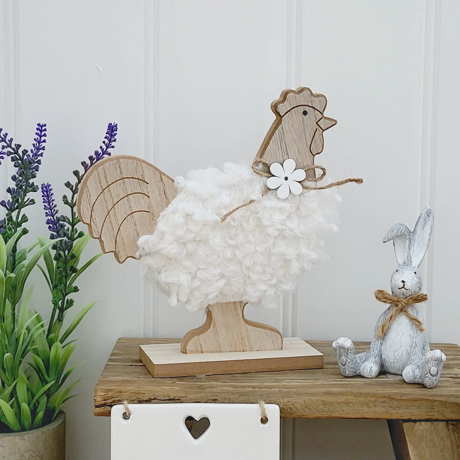 A freestanding wooden chicken ornament with a white fluffy body and a jute flower bow around neck, displayed on a wooden bench next to a grey rabbit ornament