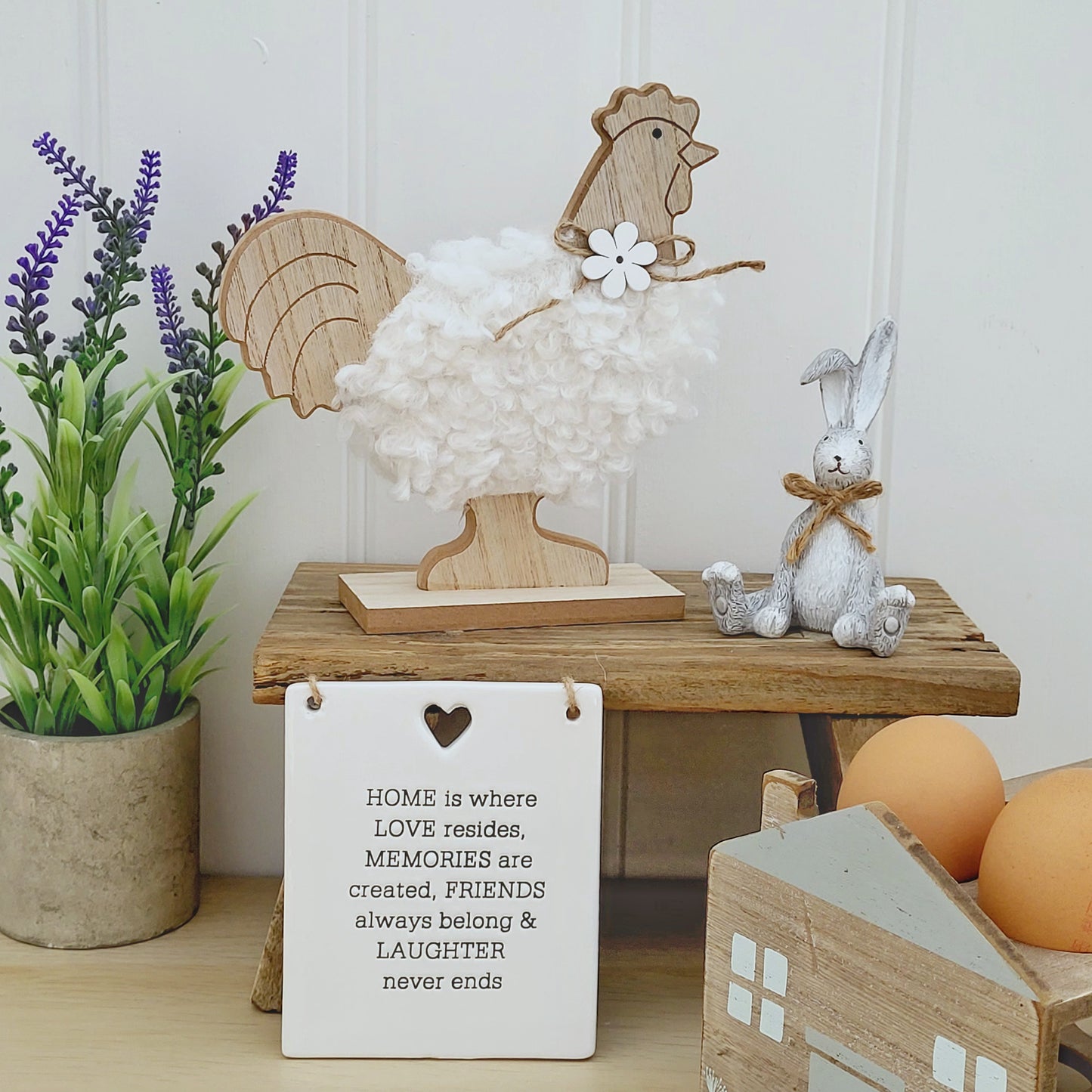 A freestanding wooden chicken ornament with a white fluffy body and a jute flower bow around neck, displayed on a wooden bench next to a grey rabbit in a kitchen worktop setting