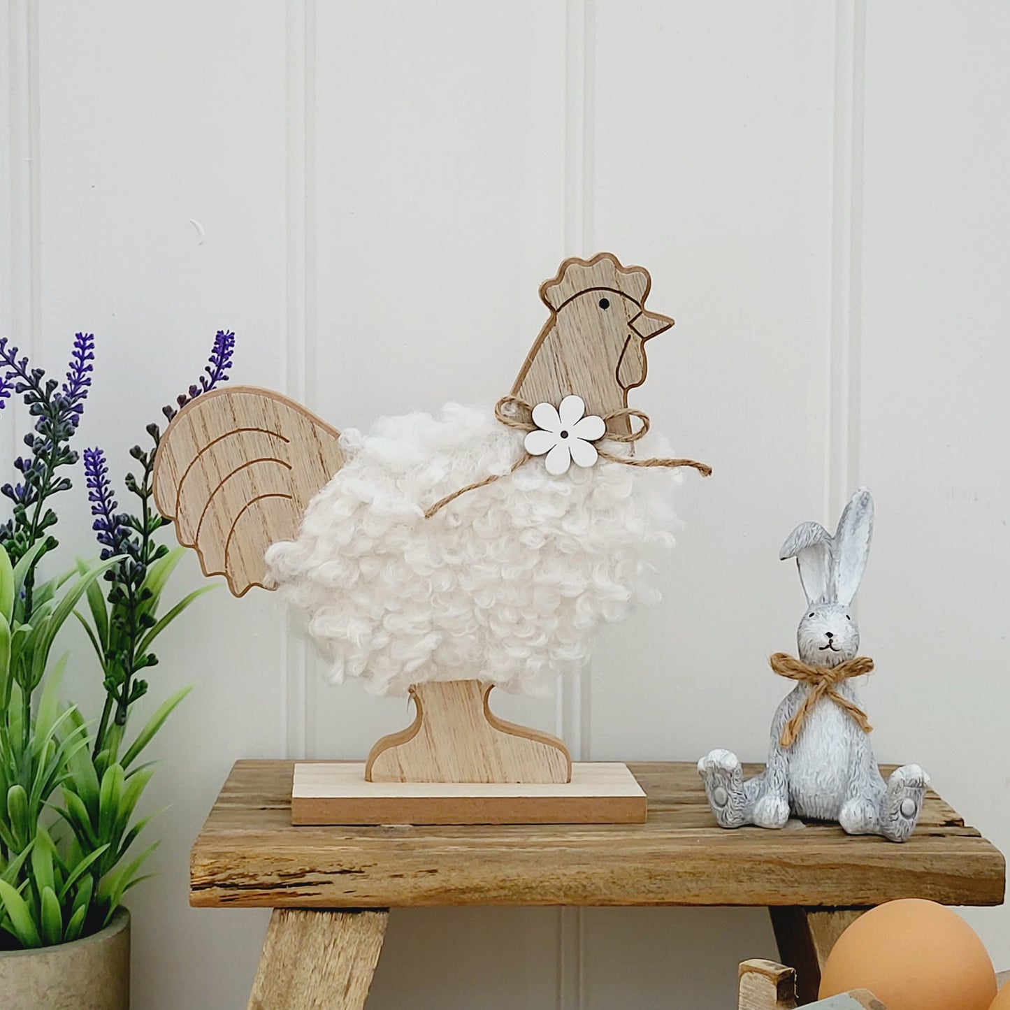 A freestanding wooden chicken ornament with a white fluffy body and a jute flower bow around neck, displayed on a wooden bench next to a grey rabbit ornament and lavender plant