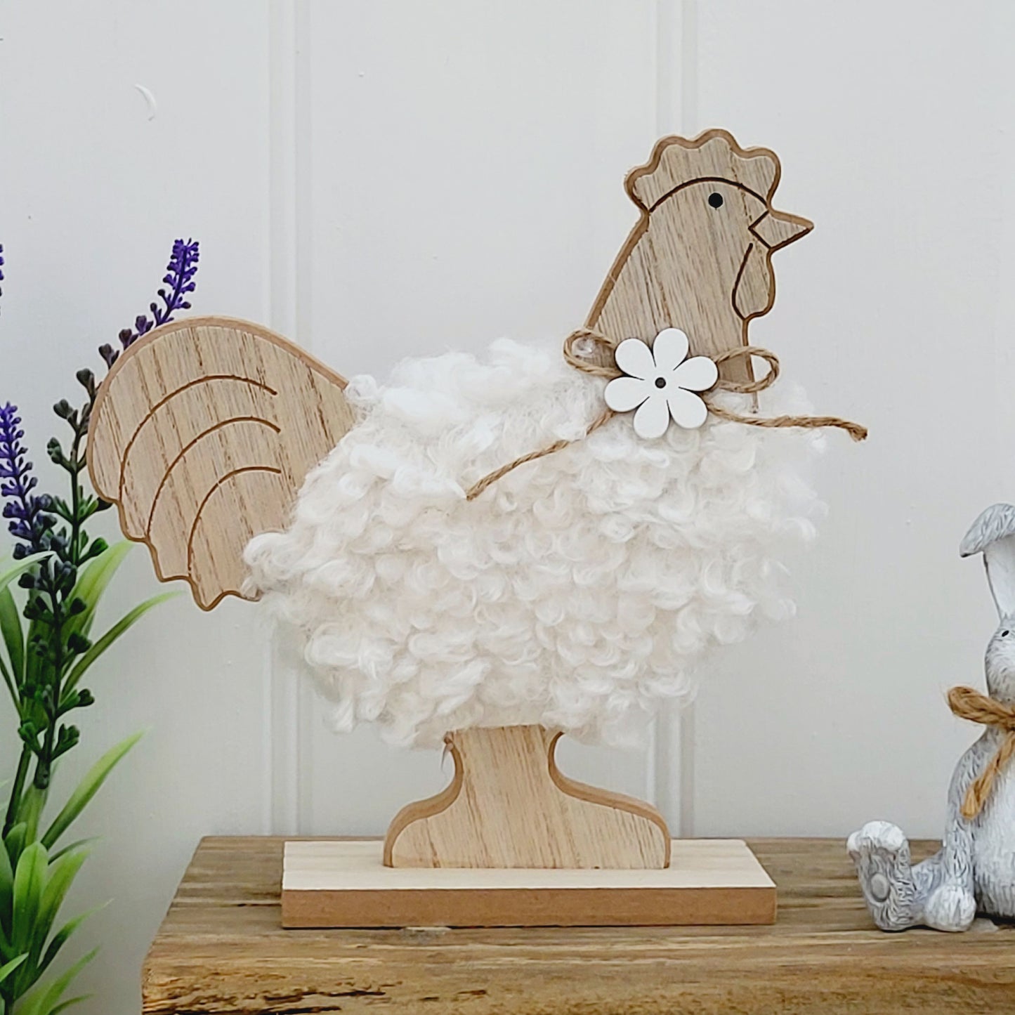 A freestanding wooden chicken ornament with a white fluffy body and a jute flower bow around neck, displayed on a wooden bench