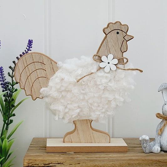 A freestanding wooden chicken ornament with a white fluffy body and a jute flower bow around neck, displayed on a wooden bench