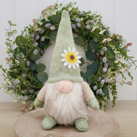 Green freestanding fabric gonk with daisy on its hat,  sat on a round wooden display board infront of a pussy willow and spring wreath greenery