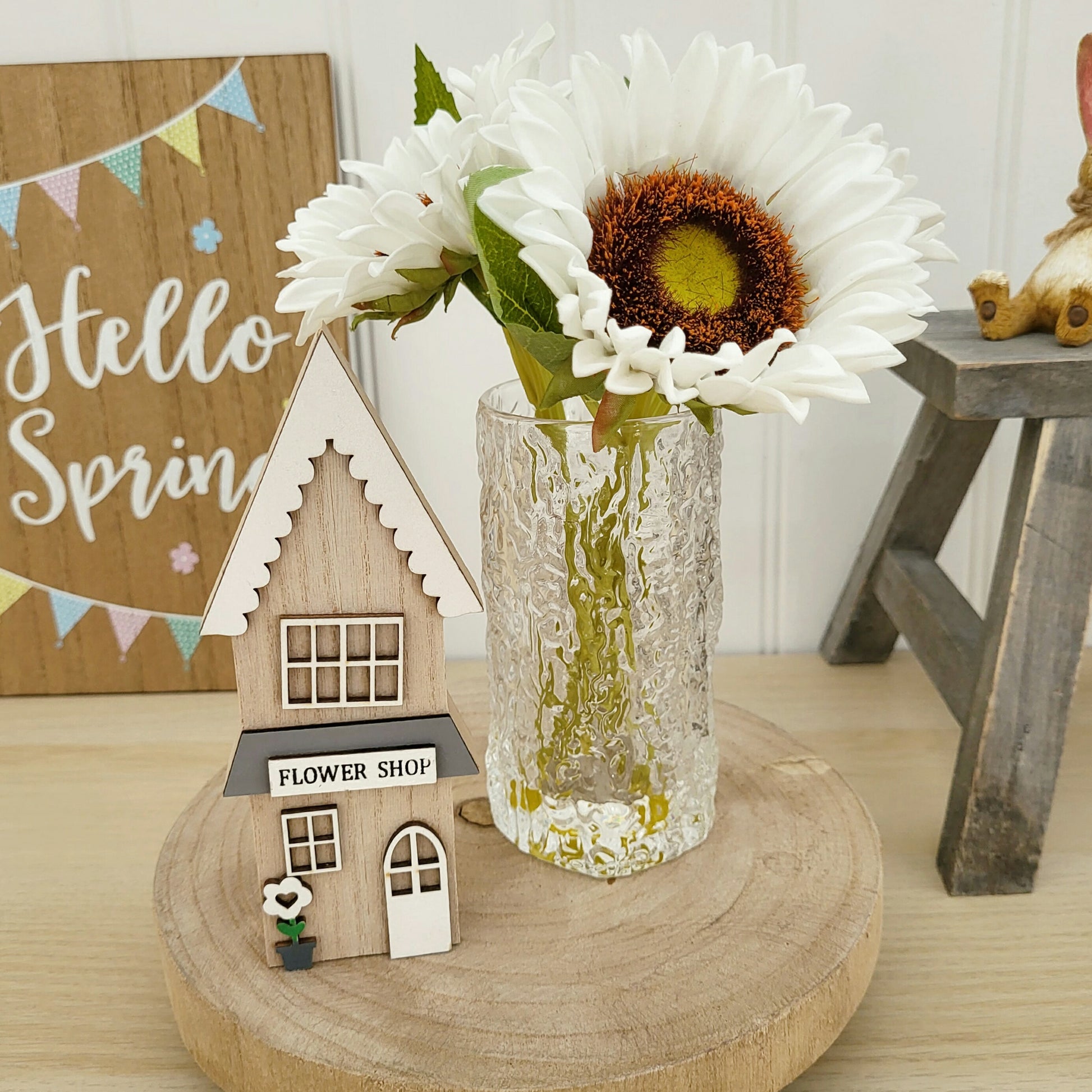 A wooden house ornament in a flower shop style displayed on a wooden board with some sunflowerd in a vase