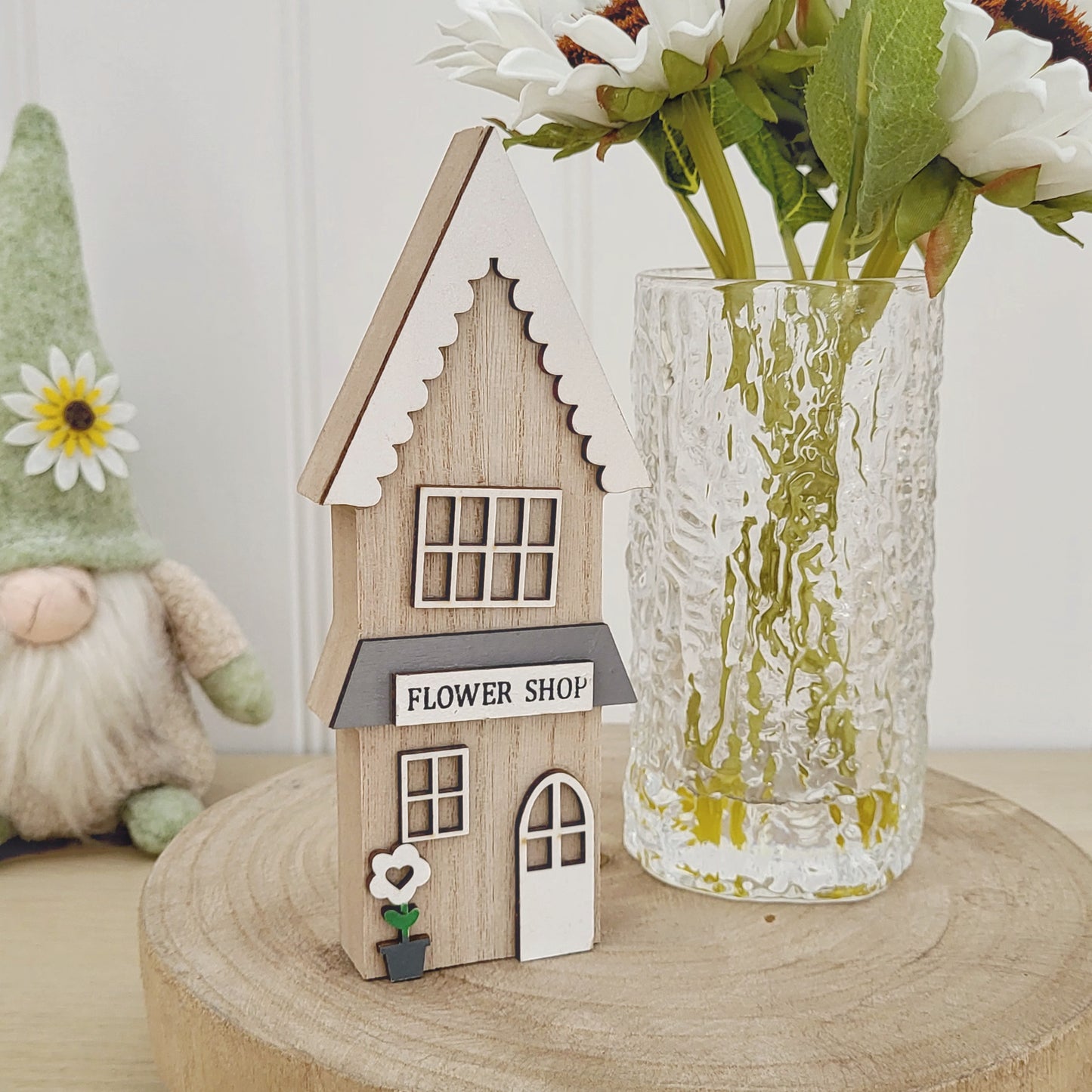 A wooden house ornament in a flower shop style displayed on a wooden board next to sunflowers in a glass vase and a sitting gonk in the background
